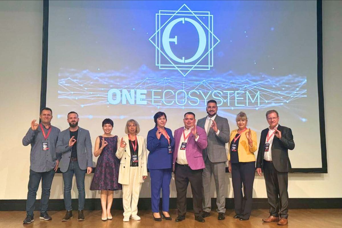 Një grup njerëzish qëndrojnë në skenë para ekranit me fjalët "One Ecosystem" të shfaqura. Ata pozojnë me të njëjtin gjest dore, duke shfaqur kohezionin dhe sinergjinë brenda komunitetit ONE ECOSYSTEM.