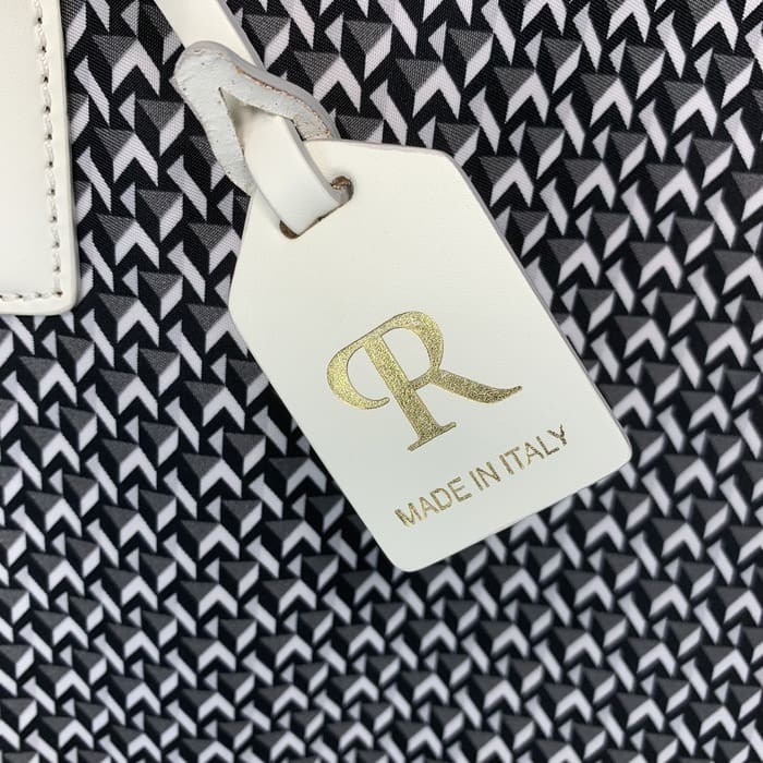 Një etiketë lëkure e bardhë ROBERTA PIERI me shkronjat "R" në ar dhe fjalët "MADE IN ITALY" varet në sfondin e një pëlhure gjeometrike bardh e zi.