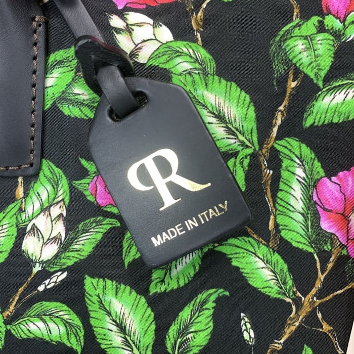 Etiketa e zezë e bagazhit ROBERTA PIERI me "R" të artë dhe tekst italian është varur nga një pëlhurë me lule me gjethe jeshile dhe lule rozë, duke plotësuar mini duffel me elegancë të përjetshme italiane.