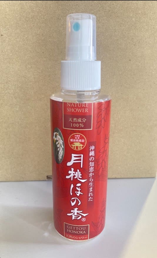 Një shishe spray e kuqe me fjalët "Nature Shower" të shkruara në një sfond bezhë. Gryka dhe kapaku i bardhë i spërkatjes.